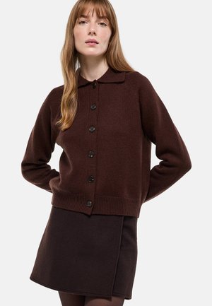 Cardigan marron en tricot avec col et fermeture à boutons, associé à une mini-jupe marron assortie, présentant une finition texturée.
