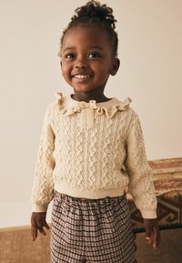 Young girl smiling, wearing a cream knitted sweater with ruffles and bow, and patterned pants, standing in a neutral-toned room.