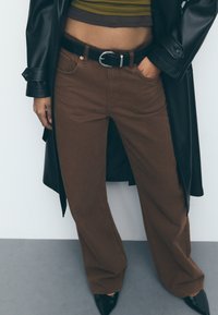 Bruine wijde broek met een zwarte riem, gedragen met een cropped gestreept top en een lange zwarte jas. Soepele stof, eenvoudige textuur.