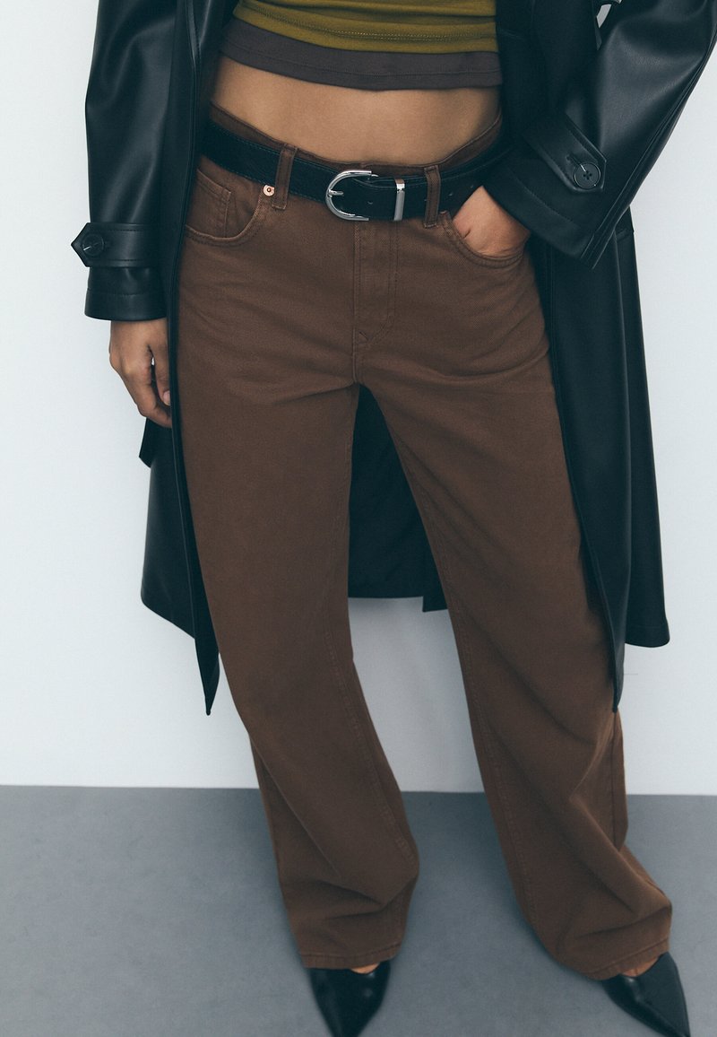 Bruine wijde broek met een zwarte riem, gedragen met een cropped gestreept top en een lange zwarte jas. Soepele stof, eenvoudige textuur.