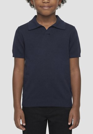 Kind draagt een donker marineblauw poloshirt met korte mouwen en een zwarte broek, staand tegen een effen lichtgrijze achtergrond.