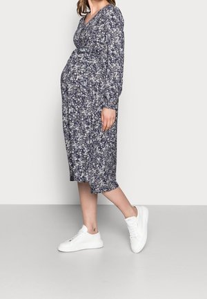 Pregnant woman wearing a long-sleeve navy floral dress and white sneakers, standing sideways against a plain gray background.