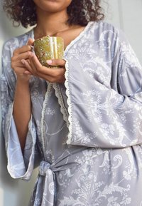 Accappatoio grigio chiaro con motivo floreale e a Vite, rifinitura in pizzo sulle maniche, legato in vita, tenuto con una tazza decorativa gialla.