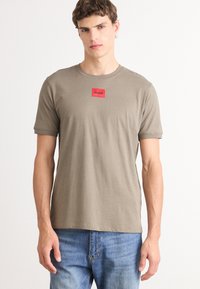 Tricou din bumbac de culoare verde măsliniu, cu guler rotund, mâneci scurte și o aplicație rectangulară roșie cu logo-ul "HUGO" pe piept.