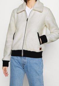 Polarowy kardigan z zipem w kolorze off-white z czarnymi akcentami, z kołnierzem, kieszeniami bocznymi oraz ściągaczami przy mankietach i u dołu.