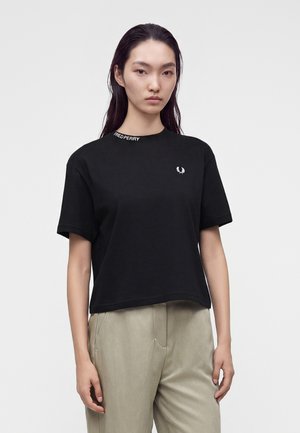 Jonge vrouw met lang donker haar, draagt een zwart Fred Perry T-shirt en lichtbeige broek, staand tegen een effen lichte achtergrond.