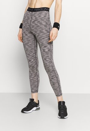 Person iført grå mønstrede Nike Pro leggings, sorte Nike håndledsbind og sorte Nike sneakers, der står foran en ensfarvet baggrund.