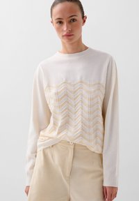 Lys cremet sweater med et chevron-mønster og strikket design i blød beige. Lange ærmer og ribbet halsudskæring; båret med beige bukser.