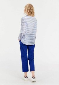Chemise blanche avec des motifs floraux bleus, associée à un pantalon bleu. Le modèle porte des baskets blanches avec des accents noirs. Texture de tissu lisse.