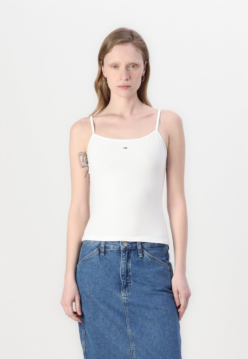 White cotton tank top with thin straps, featuring a small logo detail at the center; worn with a blue denim skirt.
