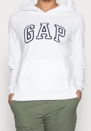 Sweat à capuche blanc avec de grandes lettres "GAP" noires contournées sur la poitrine, porté par une personne les mains dans les poches d’un pantalon vert olive.