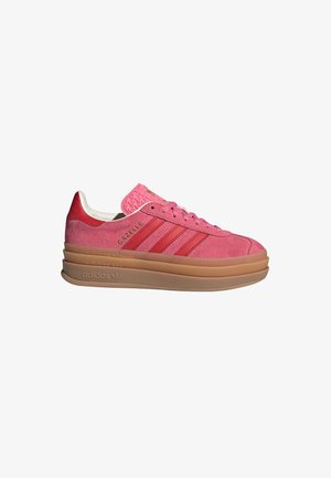 Różowy zamszowy sneaker Adidas Gazelle z czerwonymi paskami, teksturowanym językiem i grubą beżową warstwową podeszwą, widziany z boku.