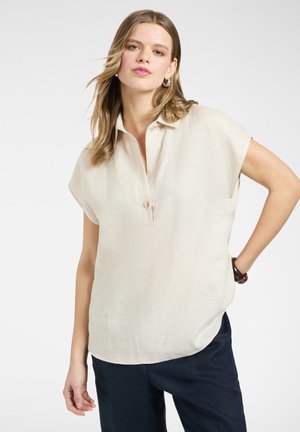 Młoda kobieta o jasnobrązowych włosach, ubrana w beżową bluzkę z krótkim rękawem i ciemne spodnie, pozująca z jedną ręką na biodrze.