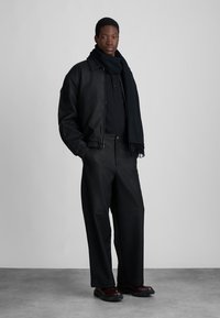 staande man in zwarte leren jas, zwarte sjaal, zwarte broek en donkerbruin schoenen tegen een effen grijze achtergrond.