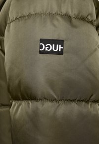 Olivenfarvet pufferjakke med kviltede sektioner og et sort rektangulært mærke, der viser brandnavnet "HUGO" i hvid skrift.