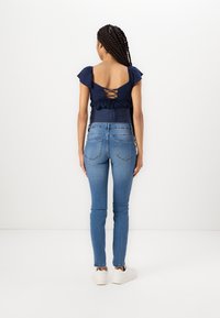 Top azul marino con mangas abullonadas y espalda con cordones. Jeans ajustados azul claro con diseño de cinco bolsillos y un ligero desgaste. Zapatillas blancas.