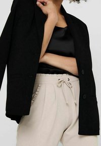 Schwarzer Blazer mit zwei Knöpfen, kombiniert mit einem schwarzen Satinoberteil und beigen elastischen Hose mit Rüschentaschen.
