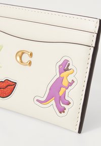 Cremefarbener Kartenhalter aus Leder, mit einem goldenen "C" und bunten Dinosaurier- und Lippenstickern. Abgerundete Kanten und minimalistisches Design.