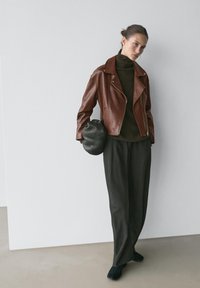 Giacca da motociclista in pelle marrone indossata sopra un dolcevita verde scuro, abbinata a pantaloni neri larghi e una borsa nera testurizzata. Design elegante e contemporaneo.