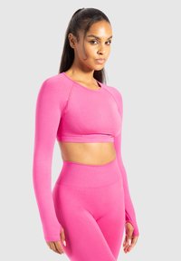 Vrouw met lang haar in een hoge paardenstaart draagt een felroze lange mouw crop top en bijpassende hoge taille leggings, poserende tegen een lichte achtergrond.