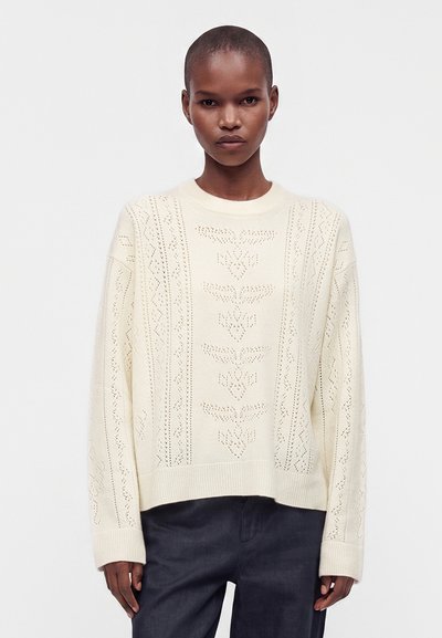 Femme aux cheveux courts portant un pull en maille crème orné de motifs floraux ajourés et un pantalon foncé sur un fond clair et uni.