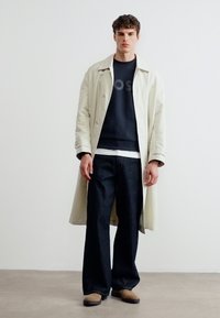 Νεαρός άνδρας που στέκεται φορώντας μπεζ παλτό τρέντσι, μαύρο φούτερ, φαρδιά σκουρόχρωμα τζιν και μπεζ παπούτσια, μπροστά σε απλό λευκό φόντο.