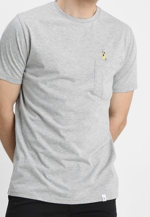 Mężczyzna w jasnoszarej koszulce z krótkim rękawem, z małym haftowanym wzorem przedstawiającym osobę na lewej kieszeni.