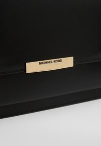 Surface en cuir noire avec une plaque métallique rectangulaire dorée gravée de "MICHAEL KORS" en lettres noires.