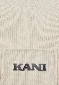 Krèmfarget strikket lue med ribbestruktur og en svart "KANI" logo-patch på den brettede bremmen.