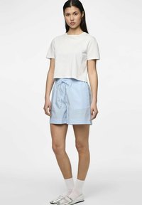 Kortärmad vit t-shirt som är avskuren, i kombination med ljusblå randiga shorts med dragsko i midjan. Modellen bär vita strumpor och silverfärgade skor.