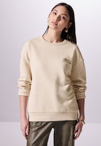 Sudadera beige con cuello redondo y puños acanalados. Presenta el texto "DESCUBRE Y EXPANDE" en color verde. Combinada con pantalones de cuero verde oliva.