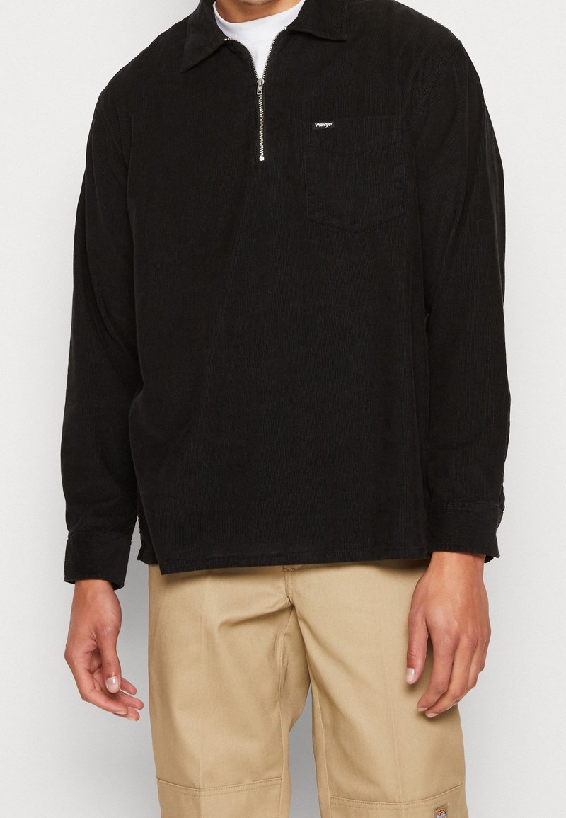 Homme portant une chemise noire à manches longues avec une fermeture éclair au quart et une poche poitrine, ainsi qu'un pantalon beige, debout sur un fond uni.