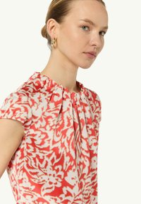 Blusa a fiori rossi e bianchi con scollatura plissettata e maniche corte, realizzata in tessuto leggero. Orecchini a cerchio dorati visibili.