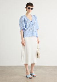 Lichtblauw kanten top met korte pofmouwen, witte midi-rok, lichtblauwe instappers en een beige schoudertas, tegen een neutrale achtergrond.