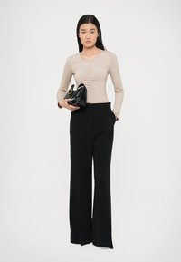 Béžový top s dlouhým rukávem a nabíráním, doplněný černými širokými kalhotami. Modelka drží také malou černou kabelku.