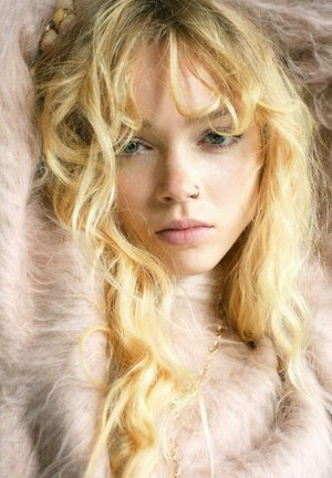 Cheveux blonds avec des boucles lâches, portant un vêtement moelleux de couleur rose pâle. Un anneau de nez notable et une délicate chaîne en or visible autour du cou.