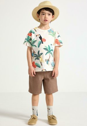 Dreng iført stråhat, t-shirt med tropisk print, brune shorts, sokker med tukan-mønster og beige sko, stående mod ensfarvet baggrund.