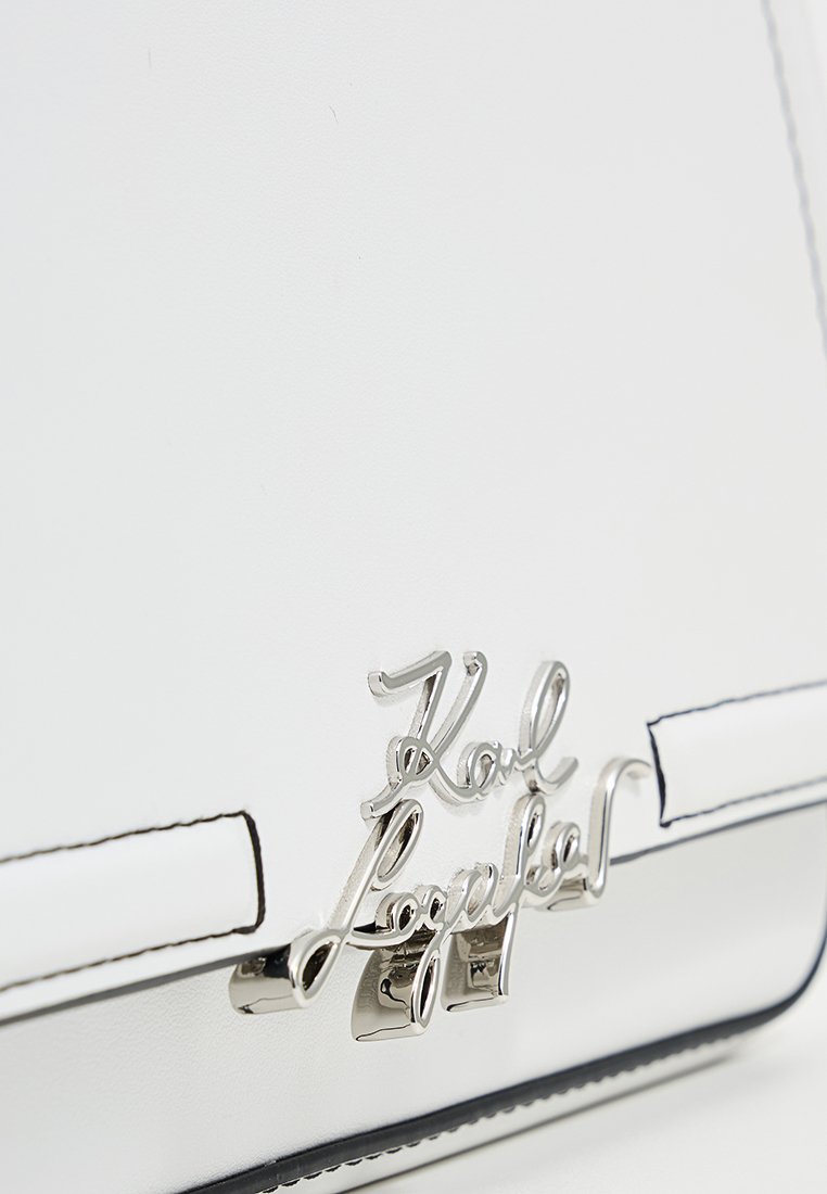 Sac à main en cuir synthétique blanc avec un design élégant, des éléments en métal argenté et un logo en relief bien visible à l'avant. Accents en bordure noire.