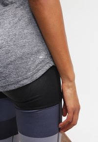 Bras droit le long du corps, portant un t-shirt Climalite gris et un legging noir et bleu à blocs de couleurs sur fond blanc.