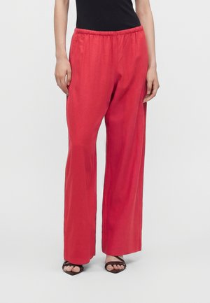 Personne portant un pantalon rouge ample à taille élastique et des sandales noires à brides, debout devant un fond blanc uni.