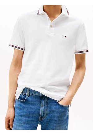 Homme portant un polo blanc à manches courtes avec un liseré rouge et noir sur le col et les manches, associé à un jean bleu, main dans la poche.