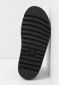 Semelle de chaussure en caoutchouc noir avec des rainures horizontales et un talon et un bout texturés, présentant un carré centré avec un symbole de croix.