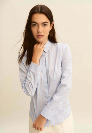 Donna con lunghi capelli scuri che indossa una camicia a righe azzurre con bottoni, che si tocca il collo, su uno sfondo beige uniforme.