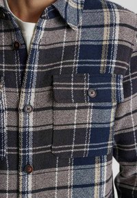 Plaid flannel shirt in navy, cream, and grey. Features two chest pockets, brown buttons, and a textured fabric weave.