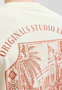 Kremkrāsas kokvilnas t-krekls ar sarkanu grafisko apdruku, kas attēlo arhitektūras elementus un textu "ORIGINALS STUDIO E." uz muguras.