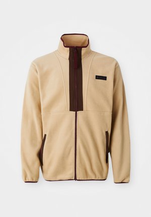 Beige fleecejack met een hoge kraag, ritssluiting aan de voorkant, bruine accenten, twee zijzakken en een klein logo label op de borst.