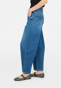 Jeans azzurri chiari a vita alta con vestibilità rilassata, caratterizzati da un design a cinque tasche e dettagli strappati; indossati con ballerine nere decorate.