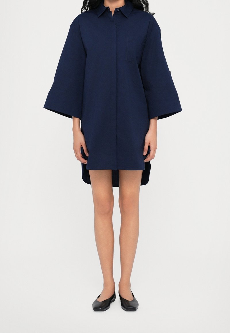Femme portant une robe chemise bleu marine oversize à larges manches 3/4 et des chaussures plates noires, debout devant un fond uni.