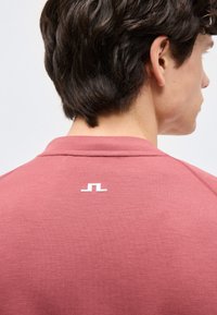 Ung person med kort mørkt hår iført en støvet rosa skjorte med en lille hvid geometrisk logo på den øverste del af ryggen.