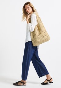 Gehaakte tas in lichtbeige met een open geweven ontwerp. Model draagt een wit langshirt, blauwe wijde broek en zwarte sandalen.